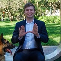 A young man in a suit jacket and white shirt holds up the University of Miami hand sign.