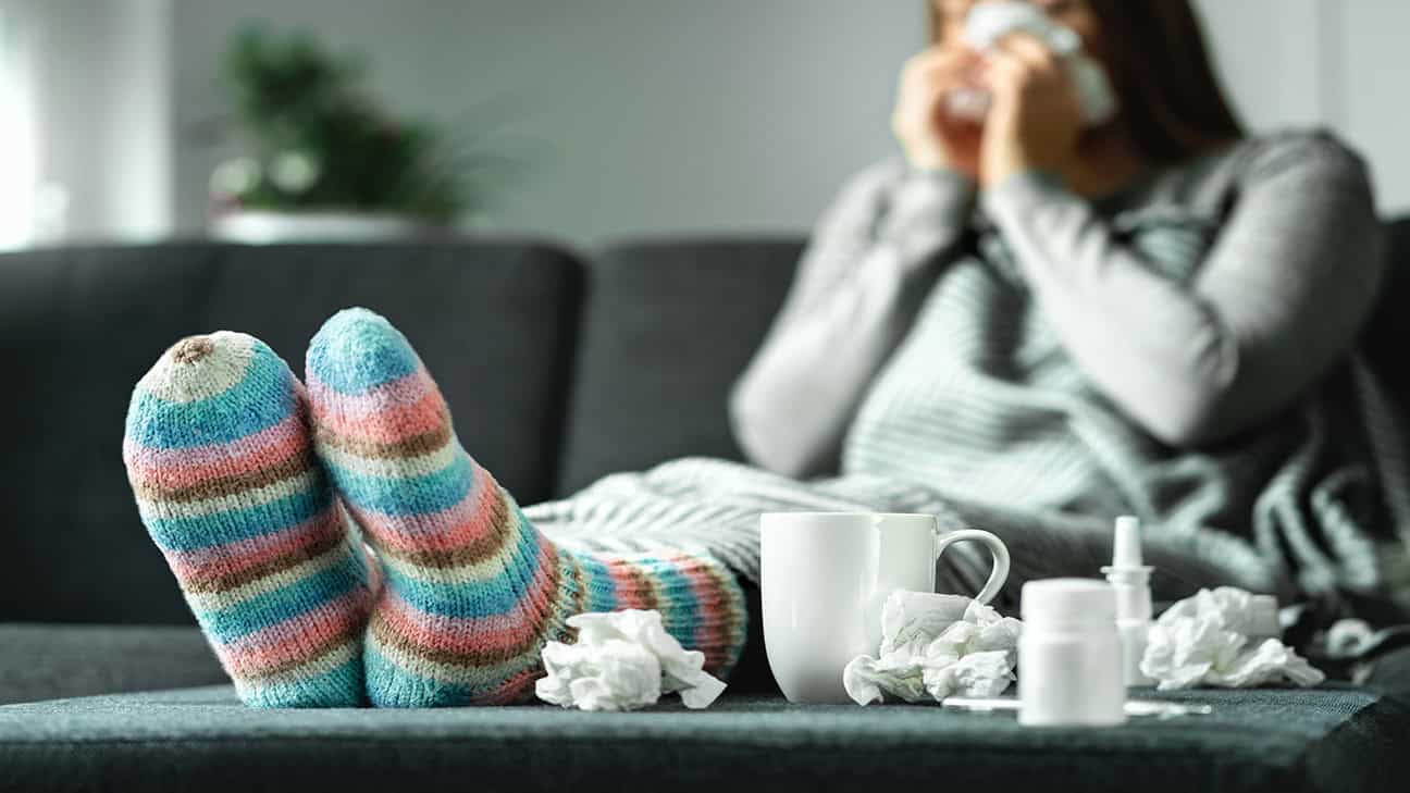 A sick woman sitting on the sofa with her feet up, blowing her nose and sneezing with tissue and handkerchief.